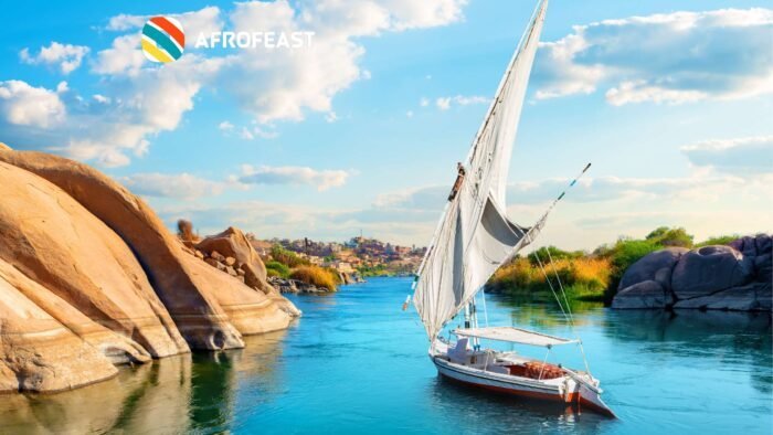 A traditional sailboat gliding on the Nile River with scenic rocky formations and a village in the background under a vibrant blue sky. The perfect Nile River Cruise example